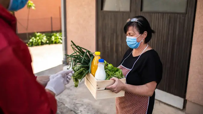 Senior woman wearing protective mask taking groceries from caring volunteer during covid-19 lockdown.