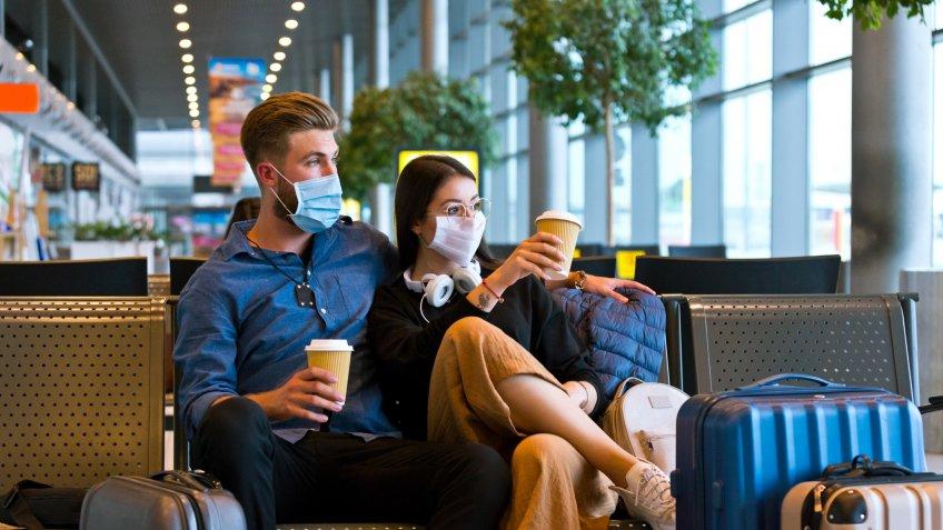 Young woman and man traveling by plane during COVID 19, wearing N95 face masks, sitting on bench with take away coffee in airport waiting area.