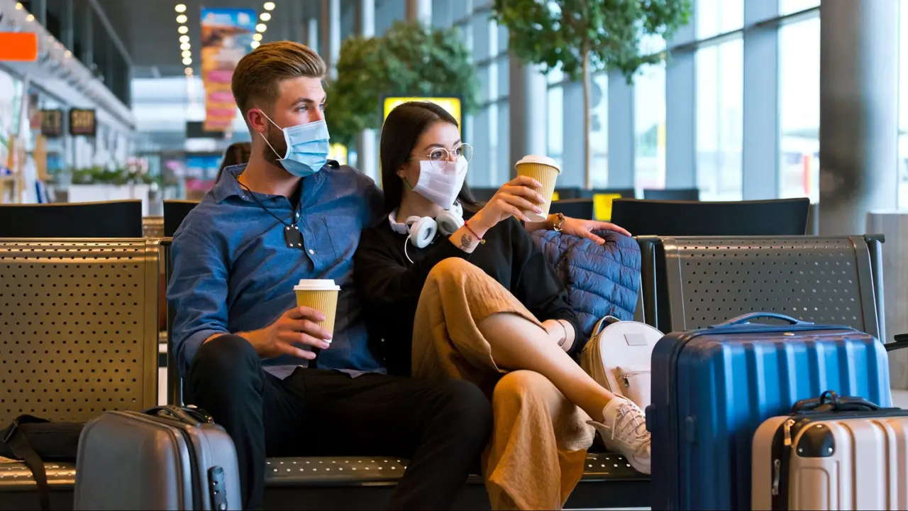 Young woman and man traveling by plane during COVID 19, wearing N95 face masks, sitting on bench with take away coffee in airport waiting area.