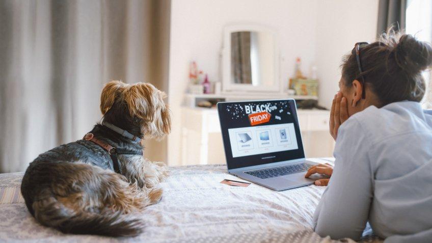Woman is buying electronics online at Black Friday while relaxing at the bed with her dog.