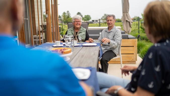two senior couples sitting together on a long table outdoors in garden enjoying afternoon coffee and sweet snacks, both sitting on one end to keep social distance in times of loosend coronavirus curfew.