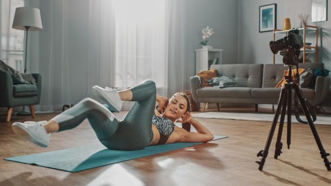 Beautiful Athletic Female Fitness Blogger is Talking on Camera and Recording her Crisscross Crunch Workout Routine in a Spacious and Bright Living Room with Cozy Modern Interior.