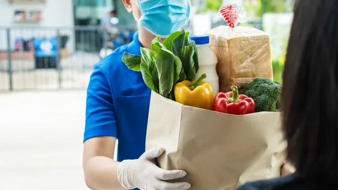 hand of food delivery service man wearing protection face mask holding fresh food set paper bag to customer at door home, express delivery, new normal, virus outbreak, takeaway food delivery concept