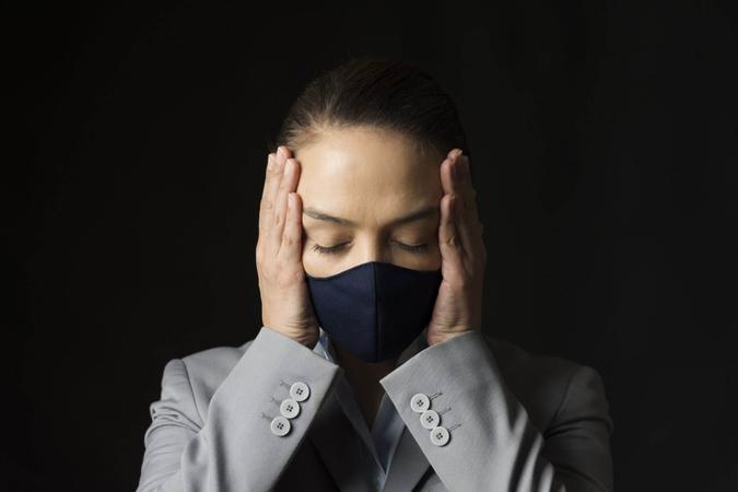Caucasian female with protective face mask and eyes closed in front of black background is holding her head with her hands.