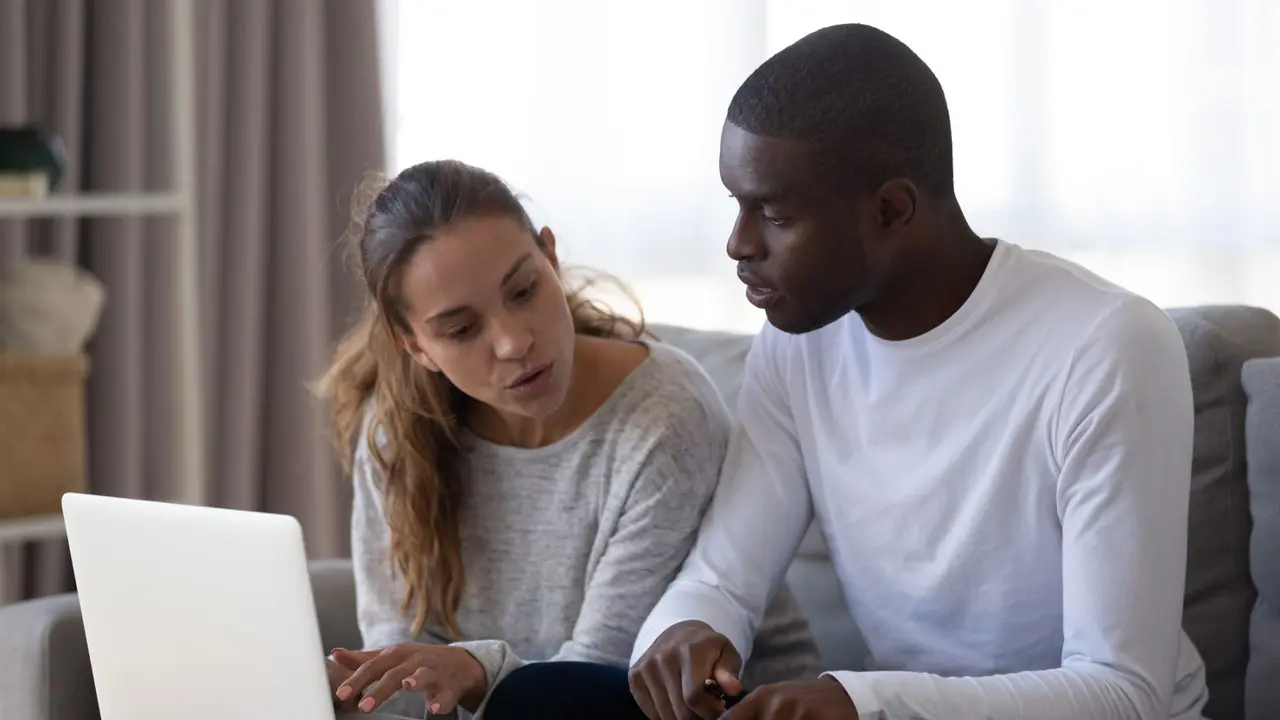 Millennial multiracial married couple siting together on couch, at table, calculating monthly family budget, shopping expenses, utility bills, using computer online banking app, submitting metrics.