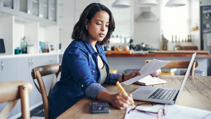 Shot of a young woman using a laptop and  going through paperwork while working from home.