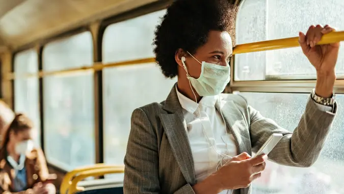 Black businesswoman with protective face mask using smart phone and looking through the window while commuting by bus.