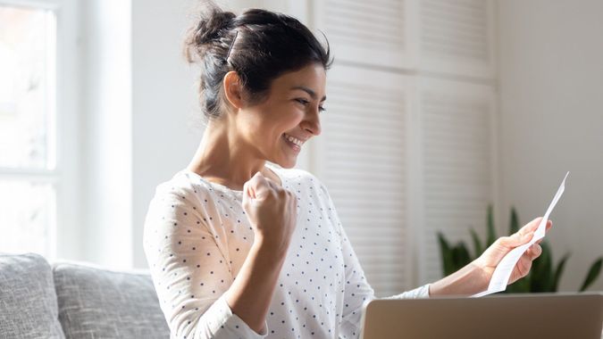 Happy Indian woman reading good news in letter, notification, holding paper, smiling girl rejoicing success, student excited by positive exam results, showing yes gesture, sitting on couch at home.