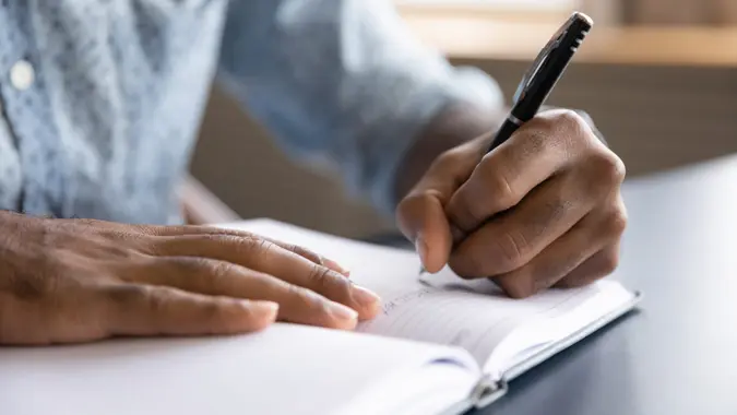 Close up view of african left-handed businessman writing in notebook, american male hands holding pen making notes planning new appointments information in organizer personal paper planner at desk.