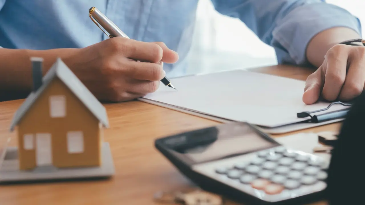 A person signing a mortgage contract of sale in a real estate office with a model house and calculator on the desk.