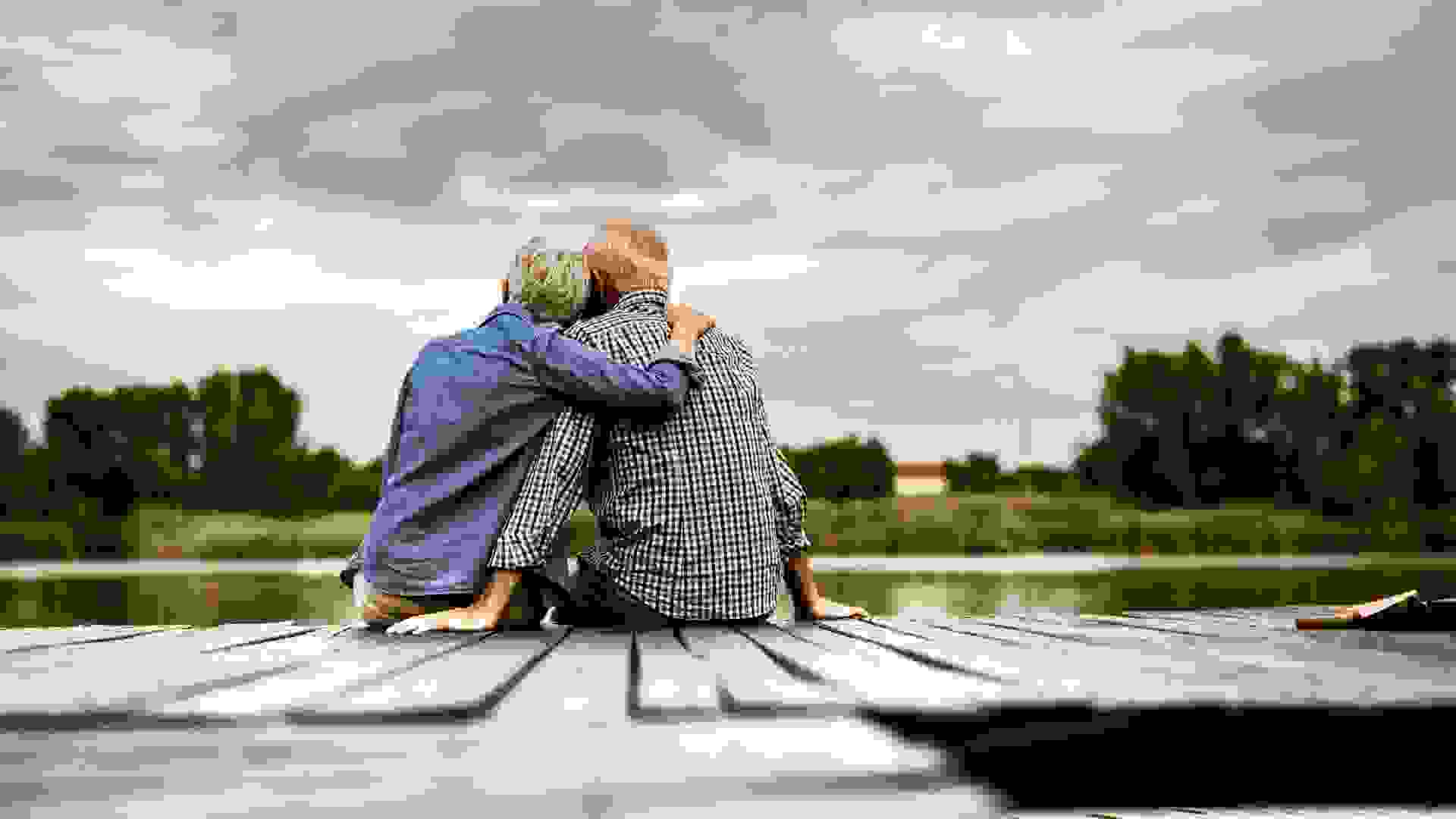 Back view of embraced mature couple looking at view while relaxing on a jetty at river.