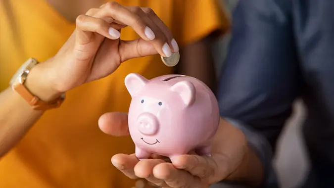 A man holds a pink piggy bank while a woman places a coin inside.