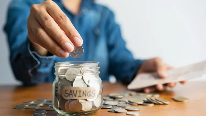 The woman hand is putting a coin in a glass  bottle and a pile of coins on a brown wooden table,Investment business, retirement, finance and saving money for future concept.