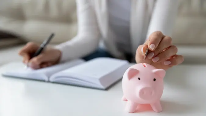 Thrifty woman sit at table hold pen writing daily expenses in diary put coin in pink piggy bank close up.