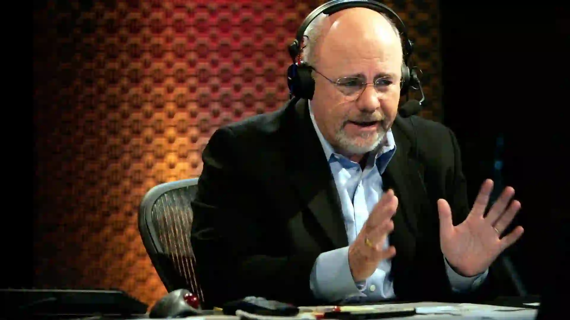 Dave Ramsey wears a headset and sits at the desk in his broadcast studio.