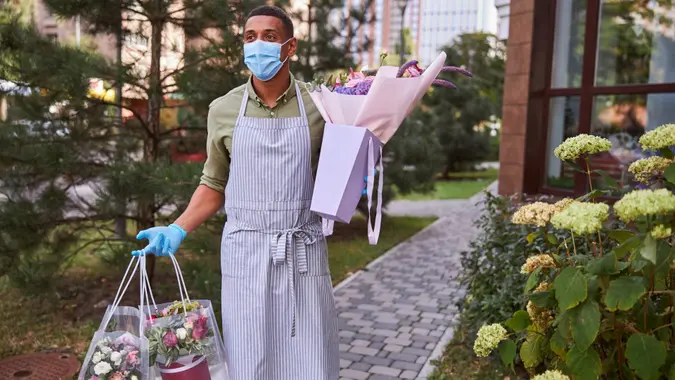 Florist in a mask walking outdoors while carrying plastic bags with flower pots and a paper package with bouquet.