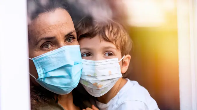 Caucasian Mature woman posing with her son, both with protective masks, very sad looking through window worried about loss of her job due Covid-19 pandemic.