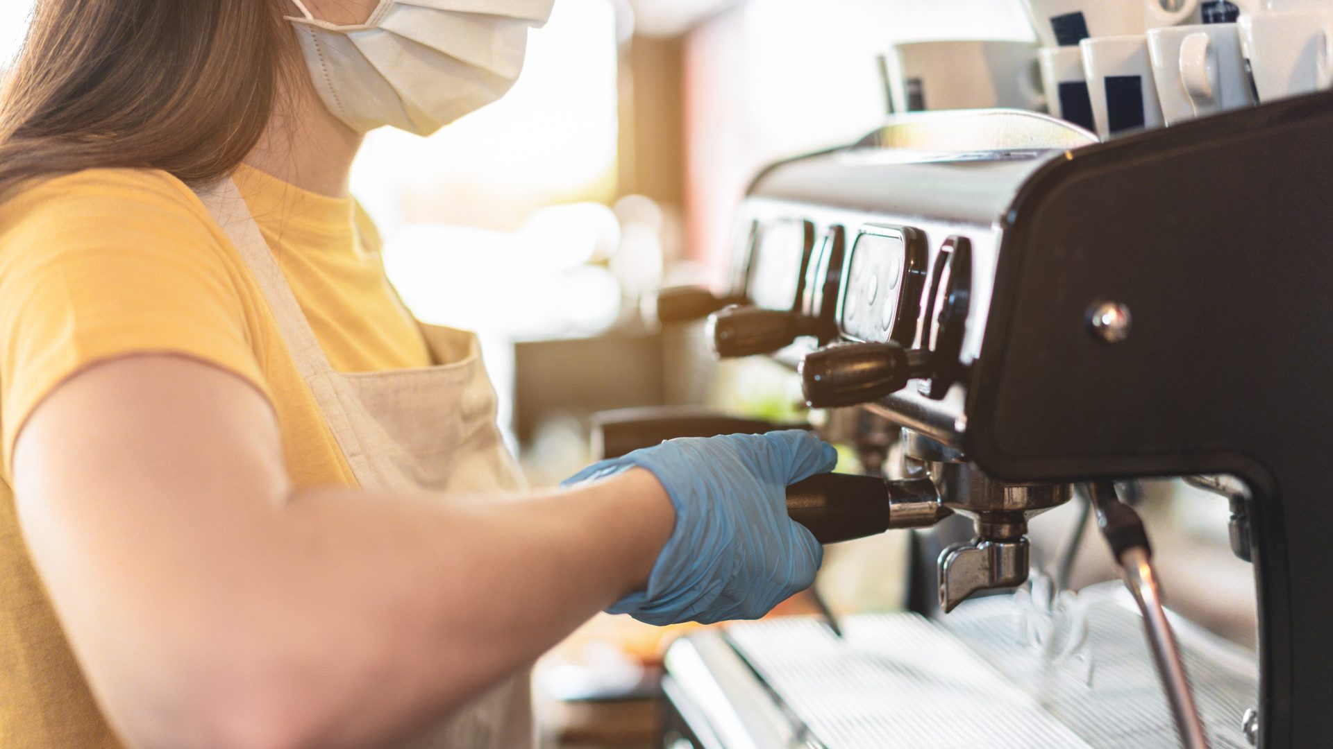 Young female working inside cafeteria bakery while making coffee wearing gloves and face mask for coronavirus spread prevention - Protective measures at work during Covid-19 outbreak - Focus on hand.