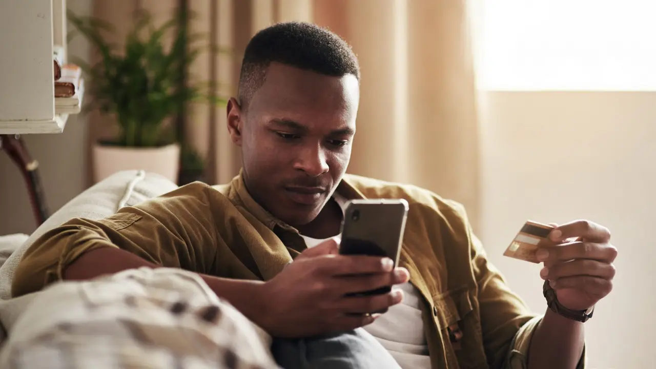 Cropped shot of a handsome young man using a smartphone and a credit card to shop online while sitting on his couch at home.