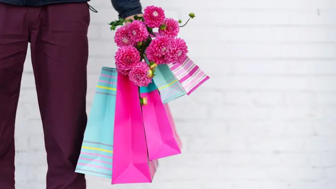 Stylish man holding flowers and pink, blue shopping bags on brick background.