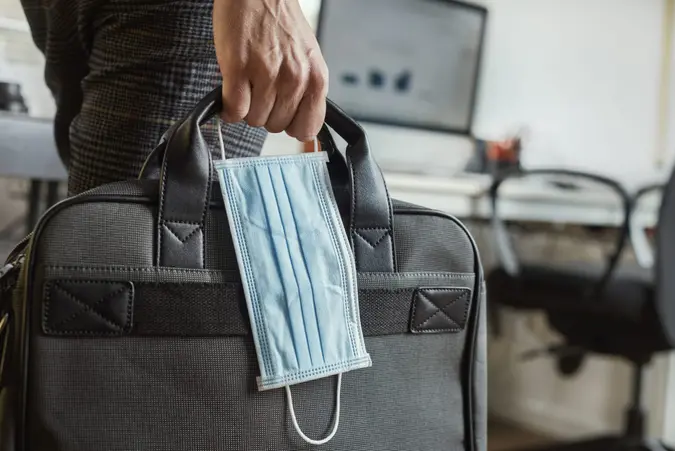 closeup of a young man in an office holding a briefcase and a surgical mask in his hand.