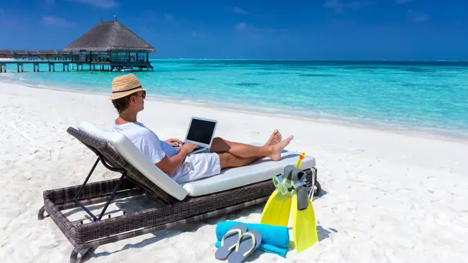 Traveler man sits on a tropical beach with his laptop and works; working in vacation concept.