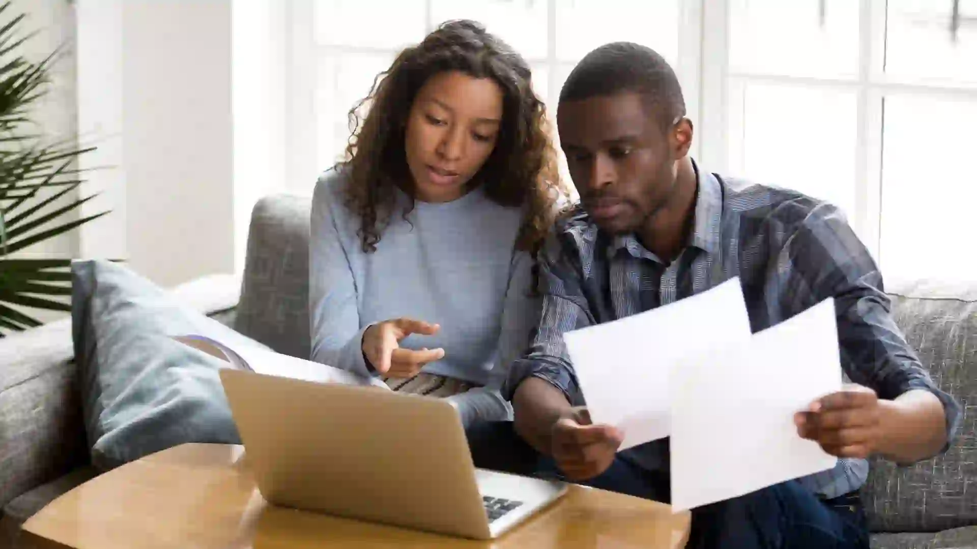 Serious African American couple reviewing documents on couch, discussing bills and bank balance.