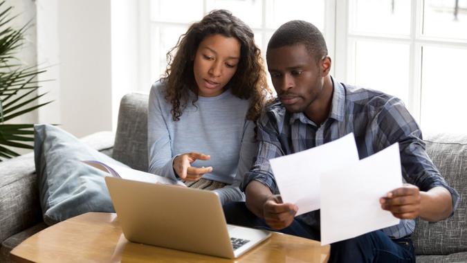Serious African American couple discussing paper documents, sitting together on couch at home, man and woman checking bills, bank account balance, terms of contract, mortgage, loan agreement.