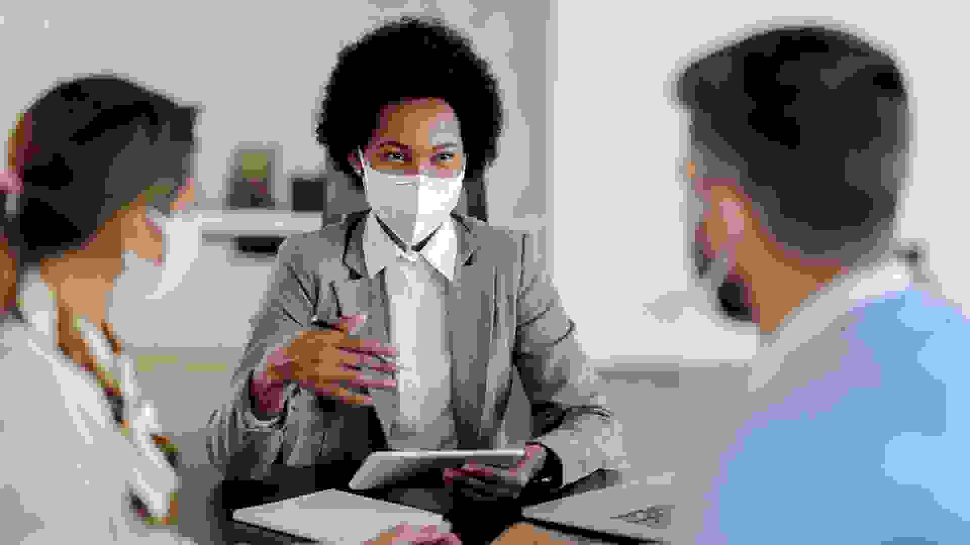 Happy black bank manager with face mask using touchpad while talking to her clients on a meeting in the office.