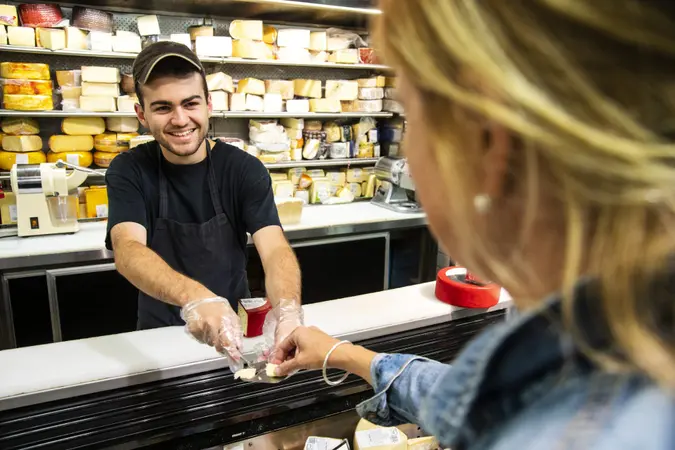 A happy young employee in the cheese section of a specialty grocery store offering a customer a sample of cheese.