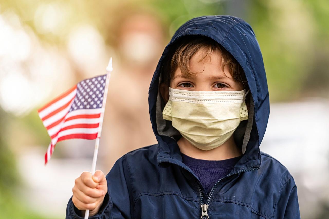 Caucasian Little boy wearing a hooded jacket, a protective mask and holding a US flag.