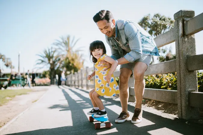 A dad helps his little girl go skateboarding, holding her waist for support.