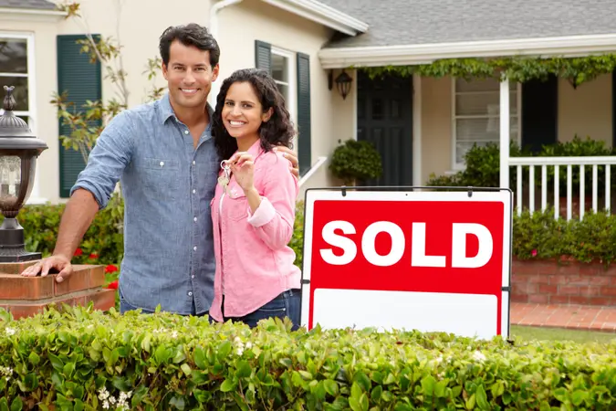 Hispanic couple outside home with sold sign holding keys in hand looking at camera smiling.