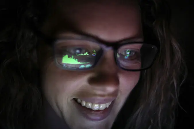 an Adult female looking at financial graphs on a laptop in a dark room with financial graphs reflected in his glasses.