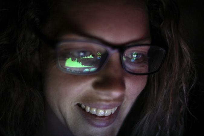 an Adult female looking at financial graphs on a laptop in a dark room with financial graphs reflected in his glasses.