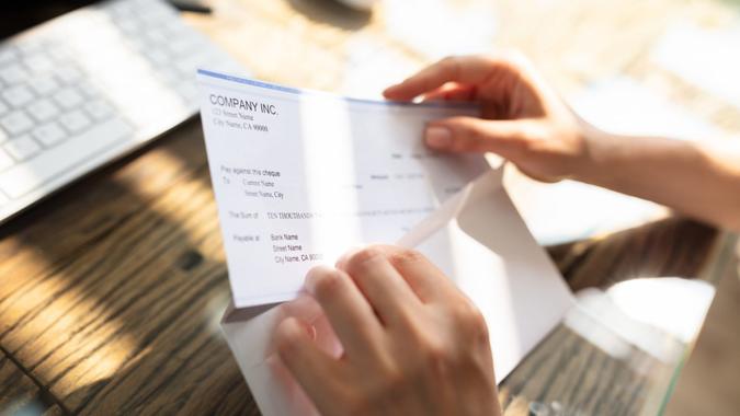 Close-up Of A Businessperson's Hand Opening Envelope With Paycheck Over Wooden Desk.