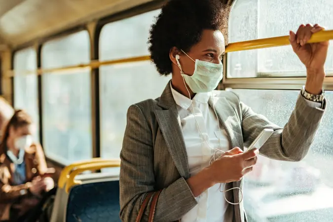 Black businesswoman with protective face mask using smart phone and looking through the window while commuting by bus.