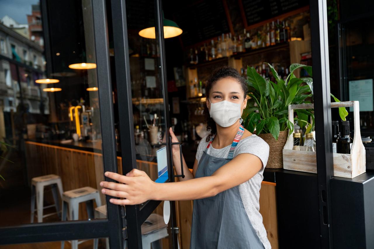 Happy business owner opening the door at a cafe wearing a facemask to avoid the spread of coronavirus â reopening after COVID-19 concepts.