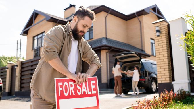 Young bearded father placing For sale sign into ground near house while putting house up for sale.