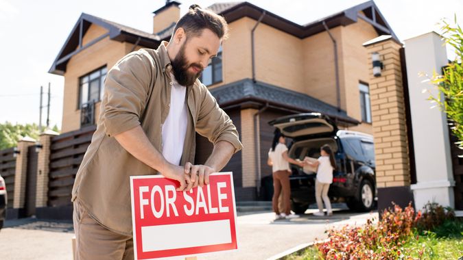 Young bearded father placing For sale sign into ground near house while putting house up for sale.