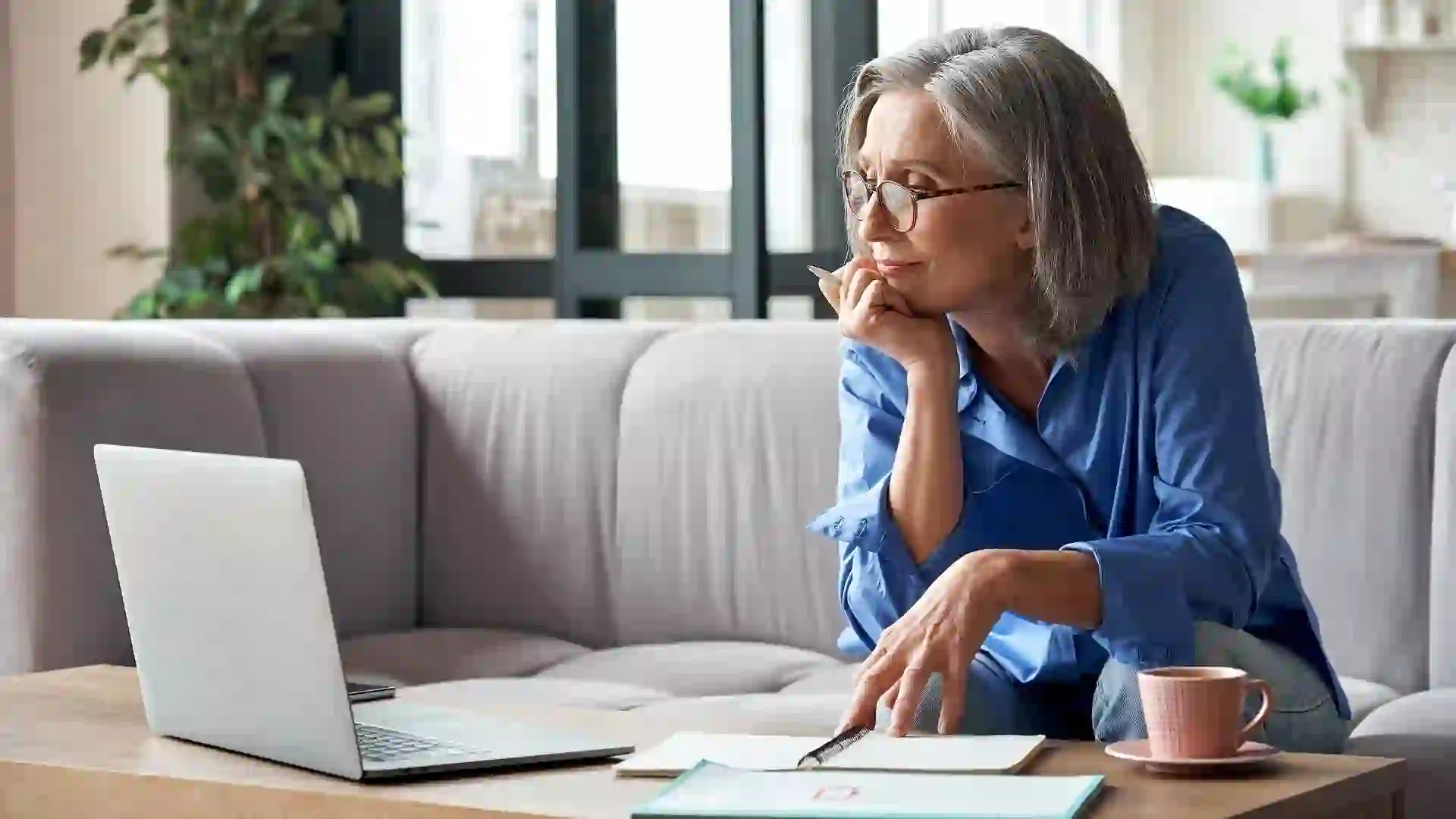 A senior woman in a blue shirt taking notes while looking at her laptop placed on the table in front of her