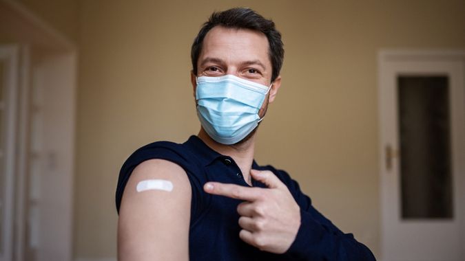 Man wearing protective face mask pointing at his arm with a bandage after receiving the covid-19 vaccine.