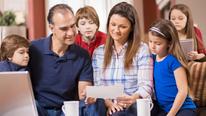 Happy family with four children discuss the family budget together as mom and dad collaborate using laptop to pay monthly bills while sitting on their living room sofa at home.