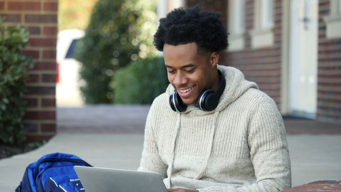  Männlicher College-Student konzentriert sich, während er einen Laptop benutzt, während er auf den Stufen eines Bildungsgebäudes sitzt und darauf wartet, dass der Unterricht beginnt.