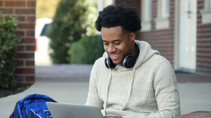 Male college student concentrates while using a laptop while sitting on the steps of an education building waiting for class to start.