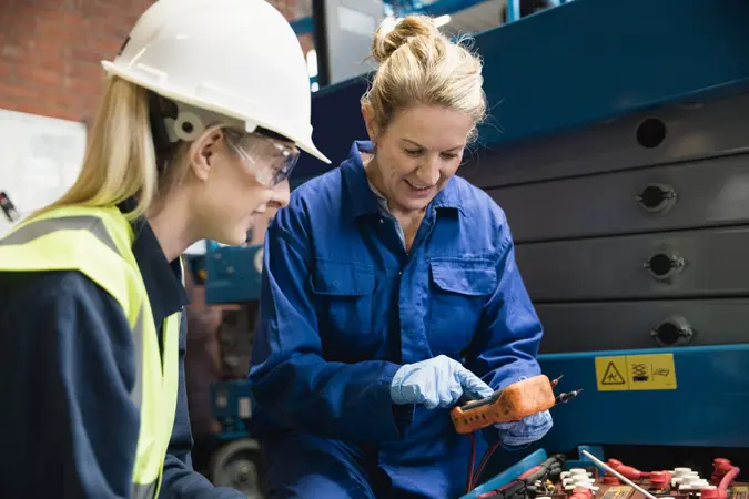 A close-up shot of two female mechanics repairing a cherry picker, one mechanic is giving a helping hand to the younger trainee.