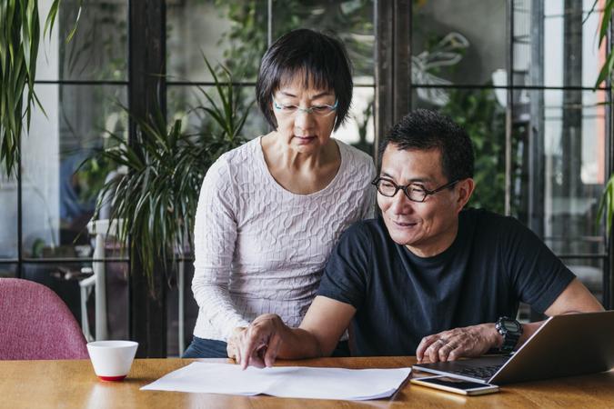 Chinese couple working on home finances with laptop, man wearing glasses and explaining, serious woman watching.