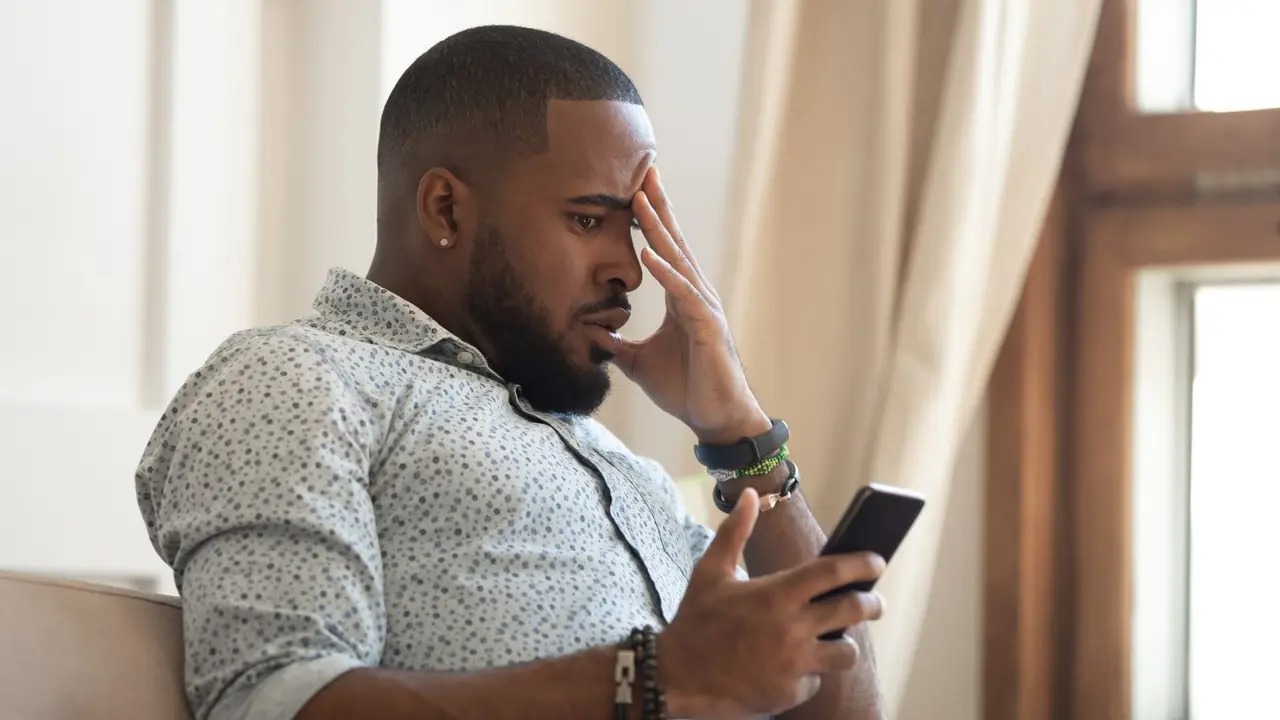 Disappointed african American millennial male sit on couch feel frustrated by unexpected bad news on cellphone, confused biracial man stressed by negative message on smartphone, cyberbullying concept.