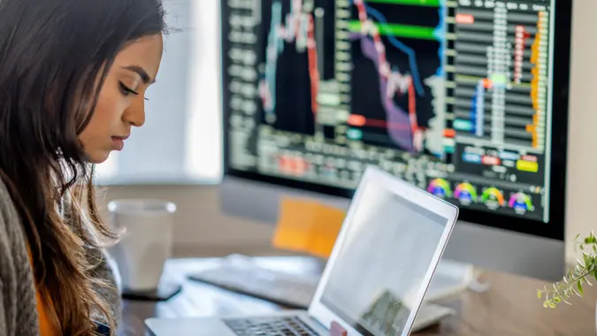 Middle Eastern woman tracking and trading stocks using laptop and desktop computer.