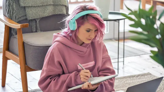 Focused hipster teen girl school college student pink hair wear headphones write notes watching webinar online video conference calling on laptop computer sit on floor working learning online at home.